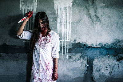 Portrait of woman with spooky make-up holding knife while standing against wall