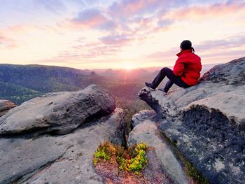 Rear view of man sitting on rock against sky during sunset