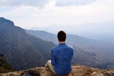 Rear view of man looking at landscape while sitting on mountain against sky