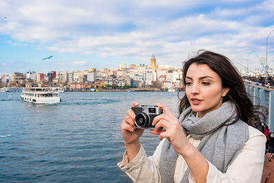 Portrait of young woman photographing by sea against sky