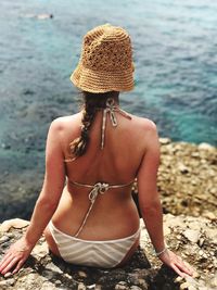 Rear view of woman in bikini sitting on rocky shore