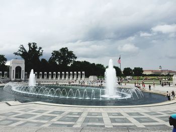 Fountain in city against cloudy sky