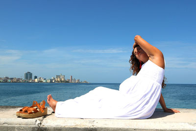 Young pregnant barefoot woman sitting on seawall against sky with her shoes next to her 