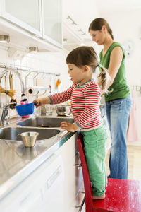 Side view of little girl filling water in container while standing on chair with mother in background at kitchen