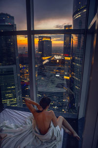 Young woman wrapped in a sheet is sitting rear on a bed in a bedroom in a skyscraper at night