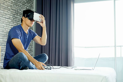 Man with virtual reality simulator playing video game on laptop while sitting in bed at home