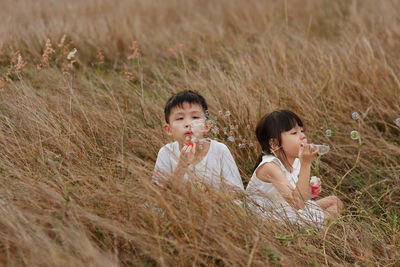 Siblings blowing bubbles while sitting on field