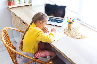 School girl in yellow sweatshirt is doing homework at home in front of laptop.