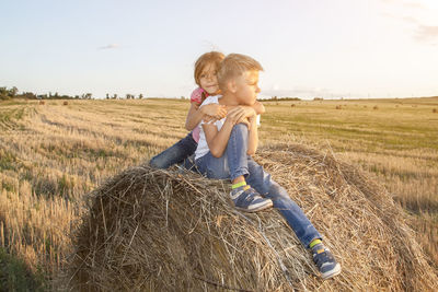 Cute sibling sitting on haystack at field against sky