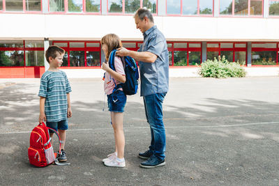 Dad escorts his children to school, helps his daughter to put on a backpack for a schoolgirl. 