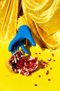 Crop anonymous person in latex glove reaching out hand to smashed ripe pomegranate with red seeds from behind shiny curtain on yellow surface in studio person