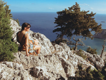 Rear view of woman sitting on rock by sea against sky