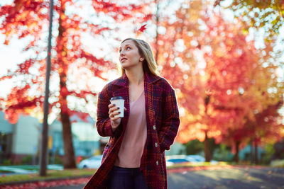 Young woman holding coffee while standing against autumn trees