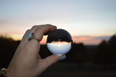 Close-up of hand holding crystal ball against sky during sunset