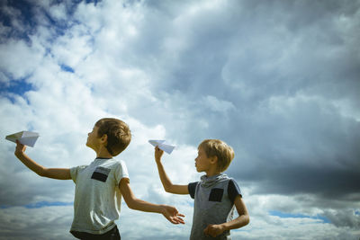 Boys playing with paper airplane against cloudy sky