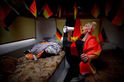 Young female soccer fan covered with german flag shouting in megaphone at home