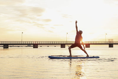 Male instructor with hand raised standing on paddleboard in sea during sunset