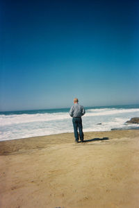 Rear view of man standing on shore at beach against clear blue sky