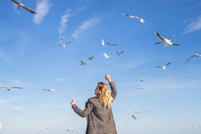 Rear view of woman standing against birds flying in blue sky