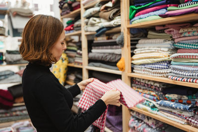 Side view of adult woman rummaging through shelf while selecting piece of cloth for work in storage room of tailor workshop