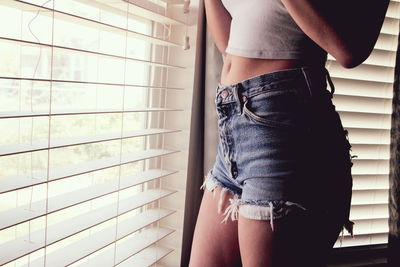 Midsection of woman wearing hot pants standing by window blinds at home