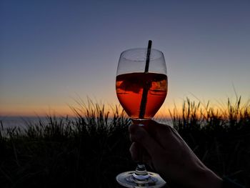 Hand holding drink against sky at sunset