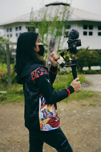 Side view of woman holding camera while standing against plants