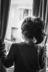 Rear view of girl looking through window while standing at home