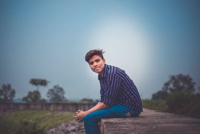 Portrait of young man sitting on retaining wall against sky
