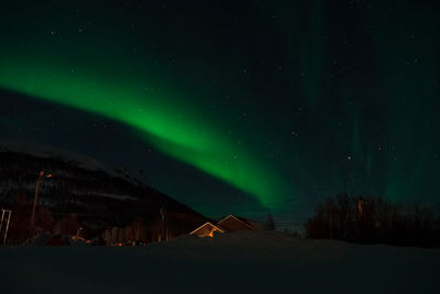 Aurora borealis against illuminated buildings and snowy mountains against the sky at night.