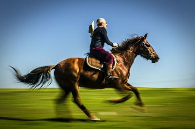 Side view of man horseback riding on field against clear blue sky