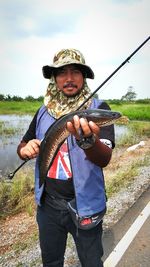 Portrait of mid adult man holding fish while standing on field against cloudy sky