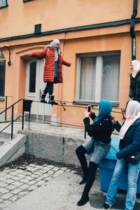 Woman with friends photographing female balancing on railing against building in city