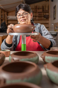 Concentrated peruvian female artisan in apron painting ceramic bowl with green color on stand while working in light professional workshop