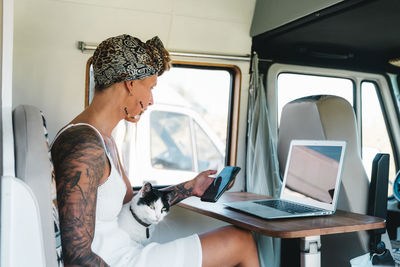Hipster female tourist with dreadlocks working on netbook and browsing on cellphone while caressing cat in caravan vehicle during road trip