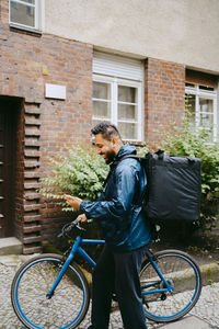 Side view of smiling male food delivery person using smart phone while standing with bicycle near building