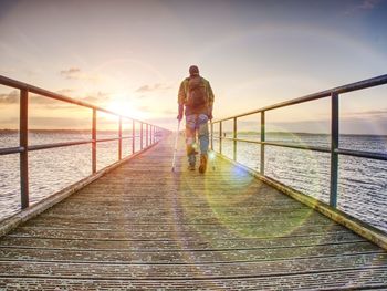 Traveler on wooden pier or jetty waiting for ferry boat by crutch. orange and blue lake morning