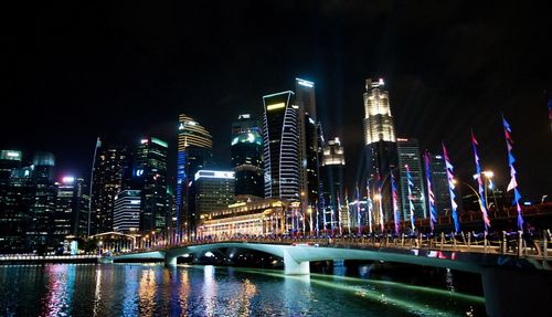 Illuminated bridge over river by buildings against sky at night
