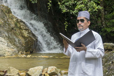 Man reading book while standing against waterfall