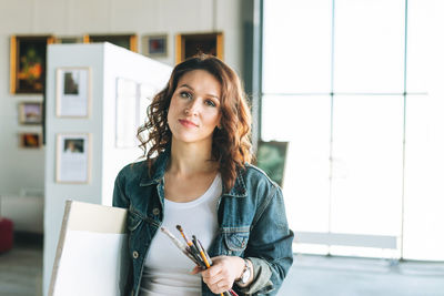 Charming young woman artist in denim jacket with paint brushes and painting on canvas 