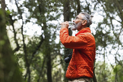 Senior man with camera drinking water while standing in forest