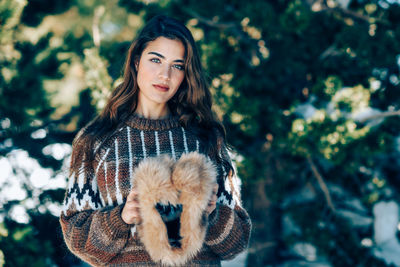 Portrait of young woman holding fur hat while standing against trees during winter