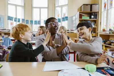 Happy teacher and boys giving high-fives while sitting at desk in classroom