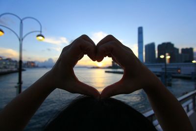 Hand holding heart shape against sky during sunset