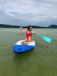 Full length of woman rowing paddleboard on sea against cloudy sky