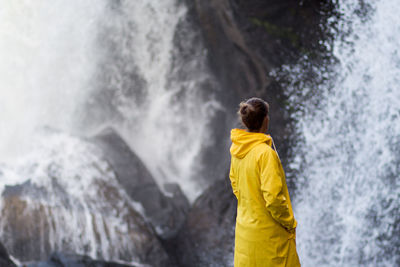 Side view of woman wearing yellow raincoat while standing against waterfall