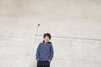 Low angle view of happy boy with hands in pockets standing against wall at recycling center