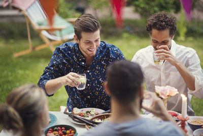 Happy men having drink while sitting with friends at table in garden party