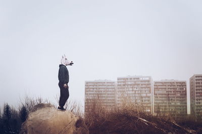 Side view of man wearing mask while standing on stone against clear sky