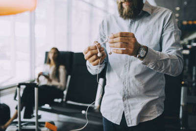 Midsection of businessman holding tangled in-ear headphones while standing at airport departure area